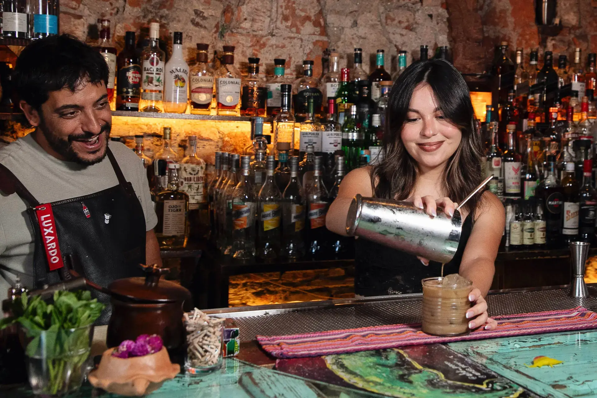 Taller de coctelería: una mujer vierte un trago y un anfitrión sonríe detrás de la barra.